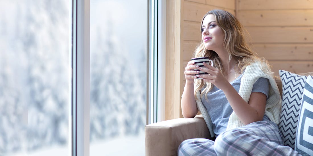 A woman is sitting on the sofa and drinking tea while looking out the window at the winter landscape