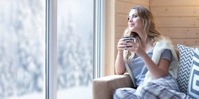 A woman is sitting on the sofa and drinking tea while looking out the window at the winter landscape