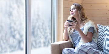 Eine Frau sitzt auf dem Sofa und trinkt Tee, während sie aus dem Fenster auf die Winterlandschaft schaut.